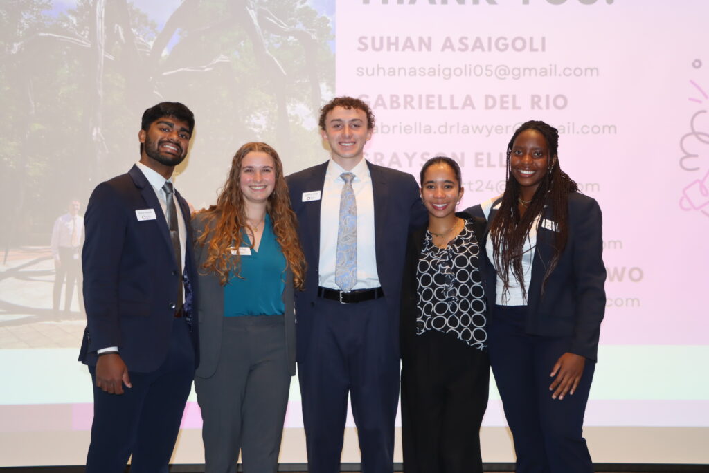 Five 2025 Morehead-Cain scholars pose together on stage for a picture. Their presentation is on a screen in the background.